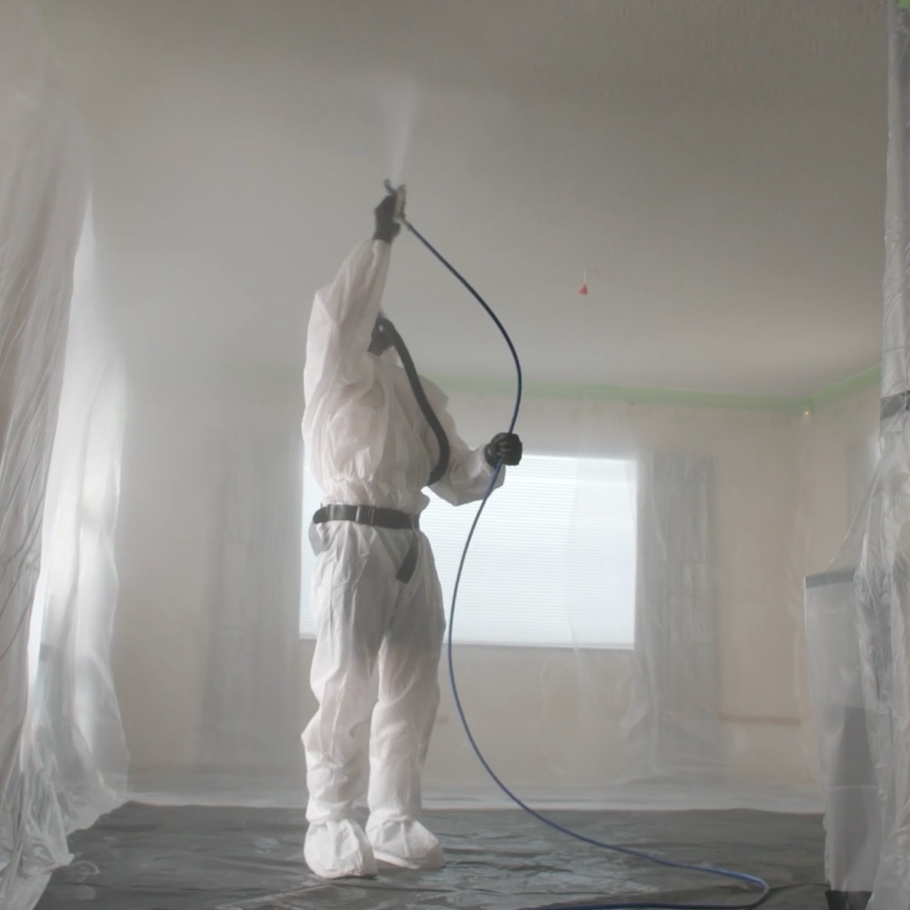 A man in a white chemical suit sprays a ceiling in an empty living room.