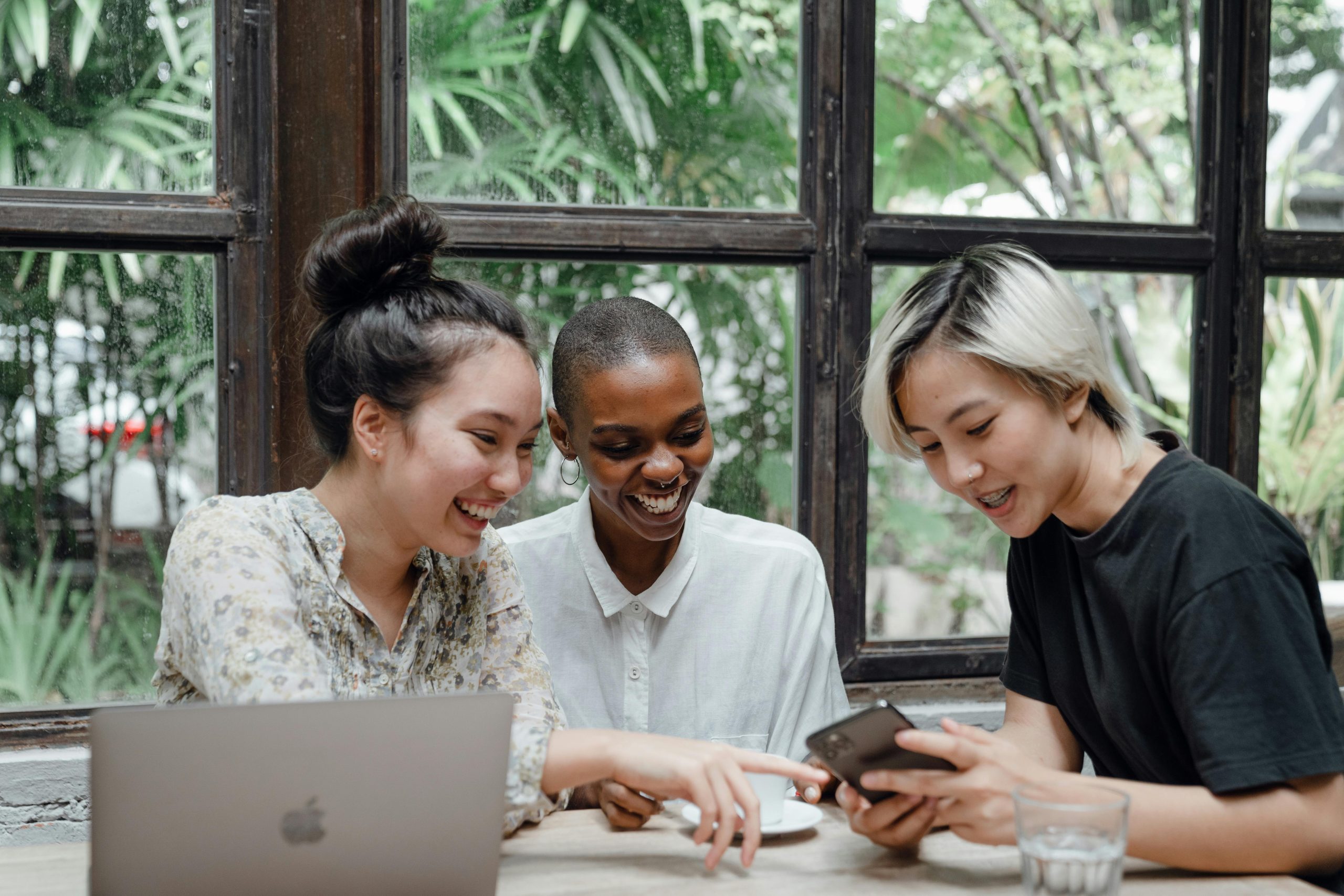 Three friends sitting at a table together and pointing and smiling at a mobile phone.